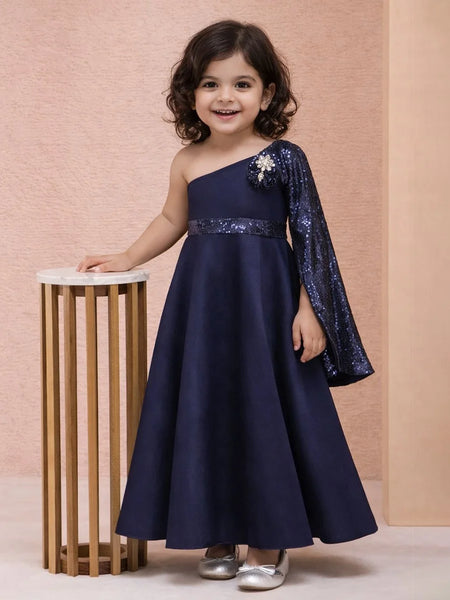 Young girl in a navy blue one-shoulder dress with sequin sleeves standing next to a small table against a pink background.