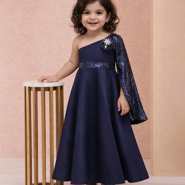 Young girl in a navy blue one-shoulder dress with sequin sleeves standing next to a small table against a pink background.