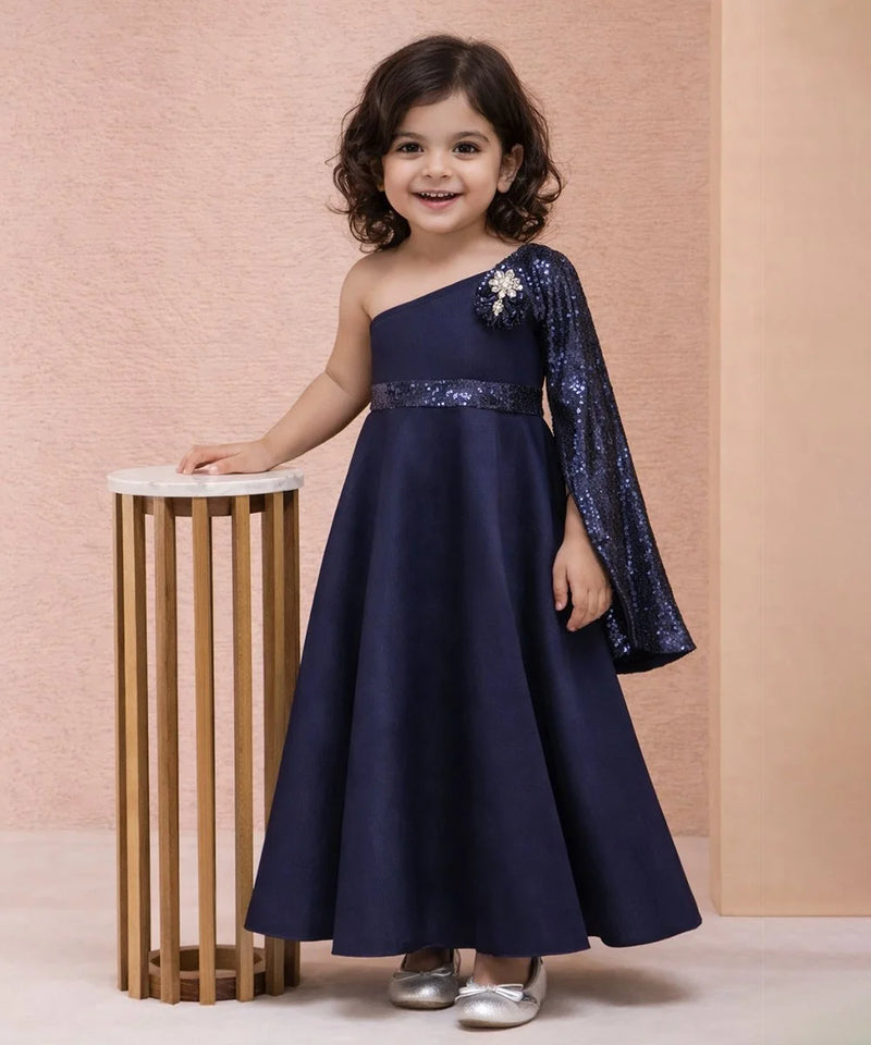 Young girl in a navy blue one-shoulder dress with sequin sleeves standing next to a small table against a pink background.