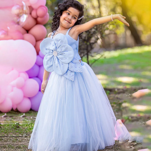 Child in a light blue dress with a large flower detail, standing outdoors with balloons in the background.