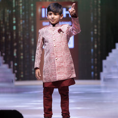 A young boy wearing a pink sherwani suit with mandarin collar and maroon-colored trousers, posing on a stage.