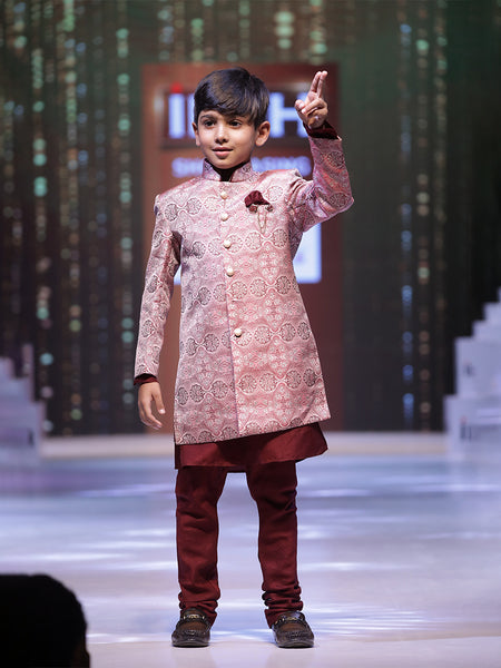A young boy wearing a pink sherwani suit with mandarin collar and maroon-colored trousers, posing on a stage.