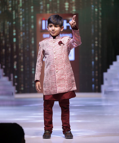 A young boy wearing a pink sherwani suit with mandarin collar and maroon-colored trousers, posing on a stage.
