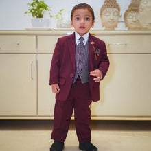 A boy wearing a maroon coat suit with a white shirt, striped tie, and matching pocket square, standing indoors.