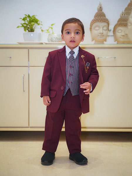 A boy wearing a maroon coat suit with a white shirt, striped tie, and matching pocket square, standing indoors.
