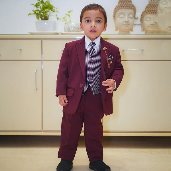 A boy wearing a maroon coat suit with a white shirt, striped tie, and matching pocket square, standing indoors.
