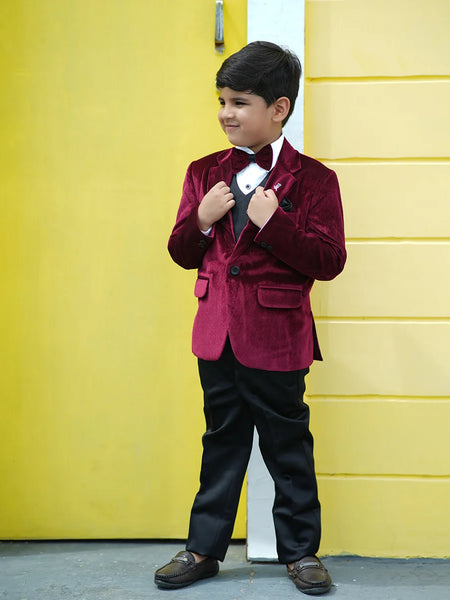A boy wearing a maroon velvet coat with a bow tie, white shirt, black waistcoat and pants, standing in front of a yellow wall.