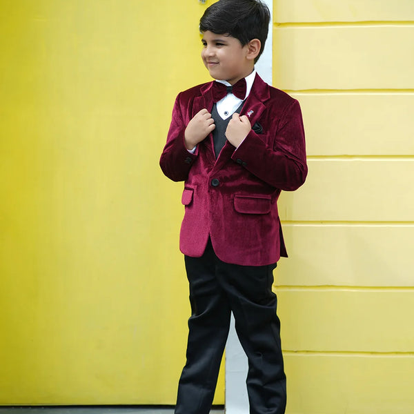 A boy wearing a maroon velvet coat with a bow tie, white shirt, black waistcoat and pants, standing in front of a yellow wall.