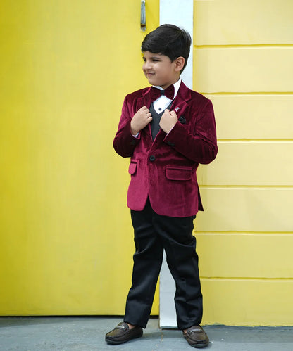 A boy wearing a maroon velvet coat with a bow tie, white shirt, black waistcoat and pants, standing in front of a yellow wall.