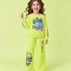 A young girl with curly brown hair wearing a bright lime green two-piece sweatshirt and pants set with colorful smiley face graphics and text, standing against a solid light green background.