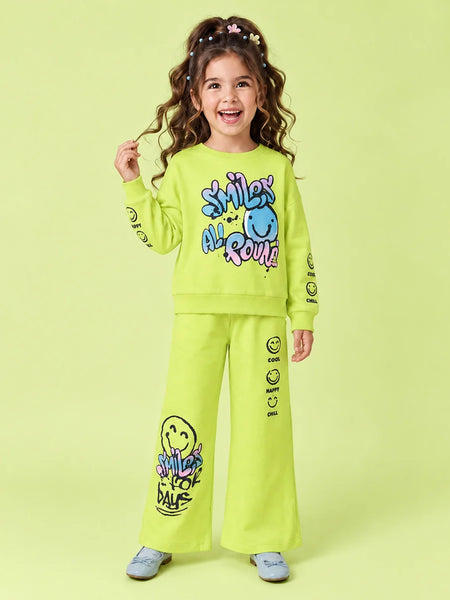 A young girl with curly brown hair wearing a bright lime green two-piece sweatshirt and pants set with colorful smiley face graphics and text, standing against a solid light green background.