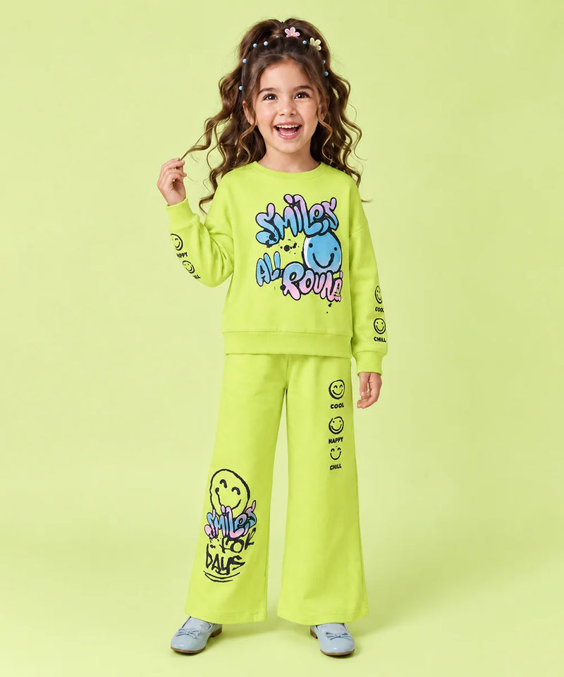 A young girl with curly brown hair wearing a bright lime green two-piece sweatshirt and pants set with colorful smiley face graphics and text, standing against a solid light green background.