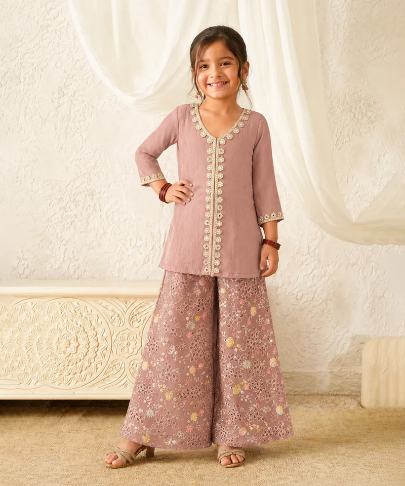 Young girl wearing a traditional outfit with a pink top and patterned pants in a decorated room.