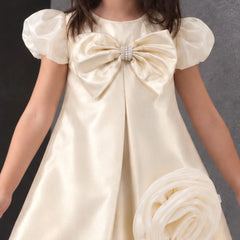 A young girl wearing a cream satin dress with a large bow at the chest, puffed short sleeves, and a matching fabric flower accessory, photographed against a dark gray background.