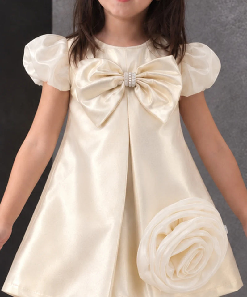 A young girl wearing a cream satin dress with a large bow at the chest, puffed short sleeves, and a matching fabric flower accessory, photographed against a dark gray background.
