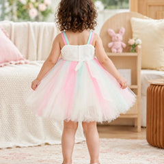 A toddler girl viewed from behind wearing a sleeveless white tutu dress with pastel pink, mint green, and light blue tulle stripes, standing in a softly decorated nursery or playroom.
