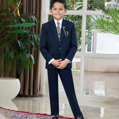A boy wearing a navy tuxedo with a white shirt, featuring a jacket, waistcoat, pants, a tie, broach, and pocket square, standing indoors.