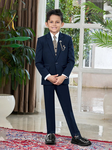 A boy wearing a navy tuxedo with a white shirt, featuring a jacket, waistcoat, pants, a tie, broach, and pocket square, standing indoors.