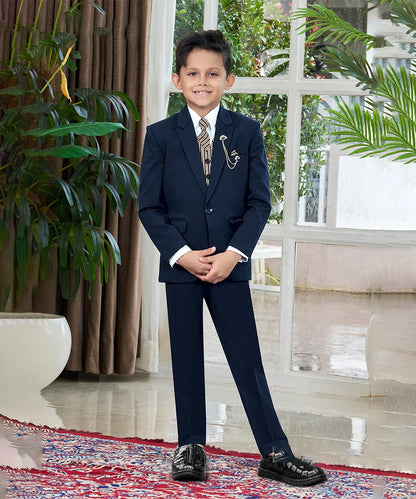 A boy wearing a navy tuxedo with a white shirt, featuring a jacket, waistcoat, pants, a tie, broach, and pocket square, standing indoors.
