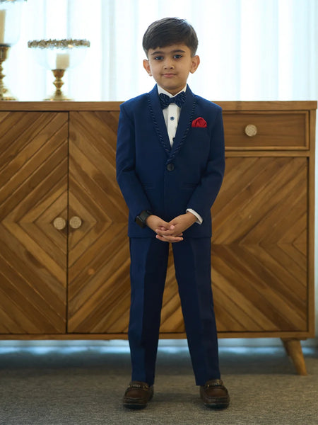 A boy wearing a navy blue tuxedo with a matching bow tie and a red pocket square, standing in front of a wooden cabinet.