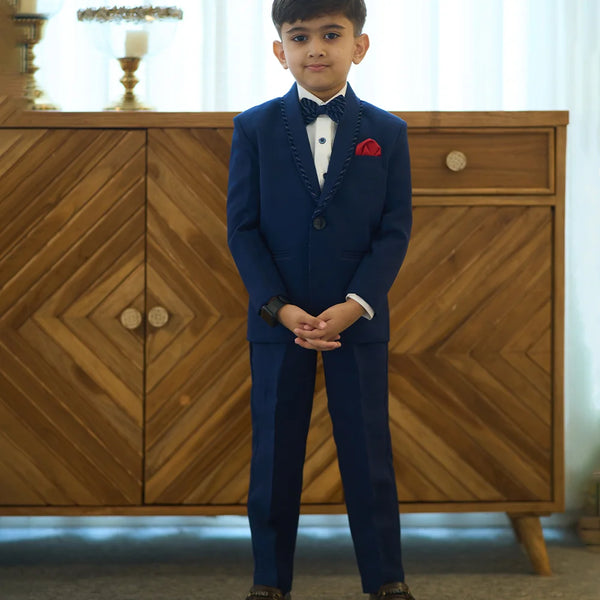 A boy wearing a navy blue tuxedo with a matching bow tie and a red pocket square, standing in front of a wooden cabinet.