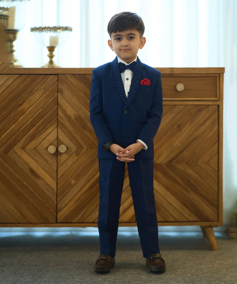 A boy wearing a navy blue tuxedo with a matching bow tie and a red pocket square, standing in front of a wooden cabinet.