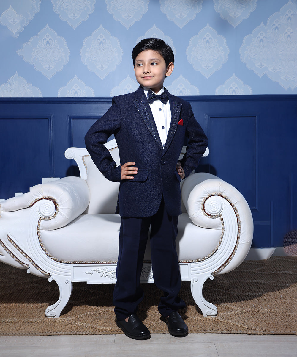 A boy aged 1-2 years in a navy blue tuxedo with a shawl collar, matching bow tie, and a pocket square standing in front of a decorative blue wall with a white chair.