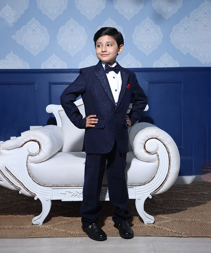 A boy aged 1-2 years in a navy blue tuxedo with a shawl collar, matching bow tie, and a pocket square standing in front of a decorative blue wall with a white chair.