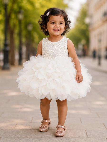 A young toddler girl wearing a sleeveless white dress with a beaded bodice and a large ruffled tulle skirt, standing on a paved outdoor walkway with trees and buildings blurred in the background.