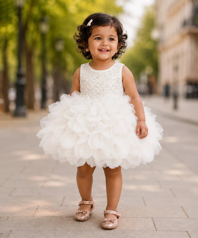 A young toddler girl wearing a sleeveless white dress with a beaded bodice and a large ruffled tulle skirt, standing on a paved outdoor walkway with trees and buildings blurred in the background.