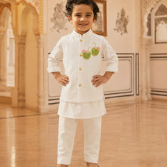 A young boy wearing a white traditional Indian outfit with gold buttons and a green embroidered motif on the chest, standing in an ornate indoor hall with beige walls and wooden floors.