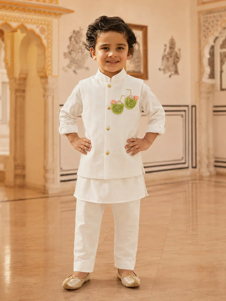 A young boy wearing a white traditional Indian outfit with gold buttons and a green embroidered motif on the chest, standing in an ornate indoor hall with beige walls and wooden floors.