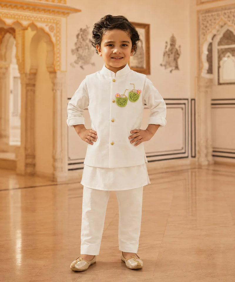 A young boy wearing a white traditional Indian outfit with gold buttons and a green embroidered motif on the chest, standing in an ornate indoor hall with beige walls and wooden floors.