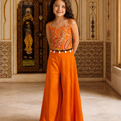 A young girl stands in an ornate indoor setting wearing a bright orange two-piece outfit with a paisley-patterned bodysuit top and matching wide-leg pants, accented by a black belt with white tassels.