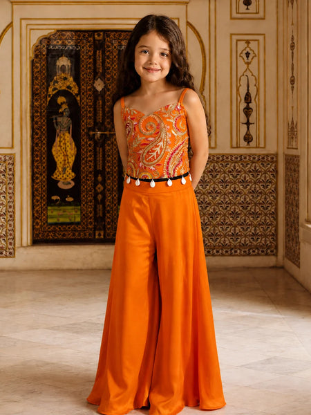 A young girl stands in an ornate indoor setting wearing a bright orange two-piece outfit with a paisley-patterned bodysuit top and matching wide-leg pants, accented by a black belt with white tassels.
