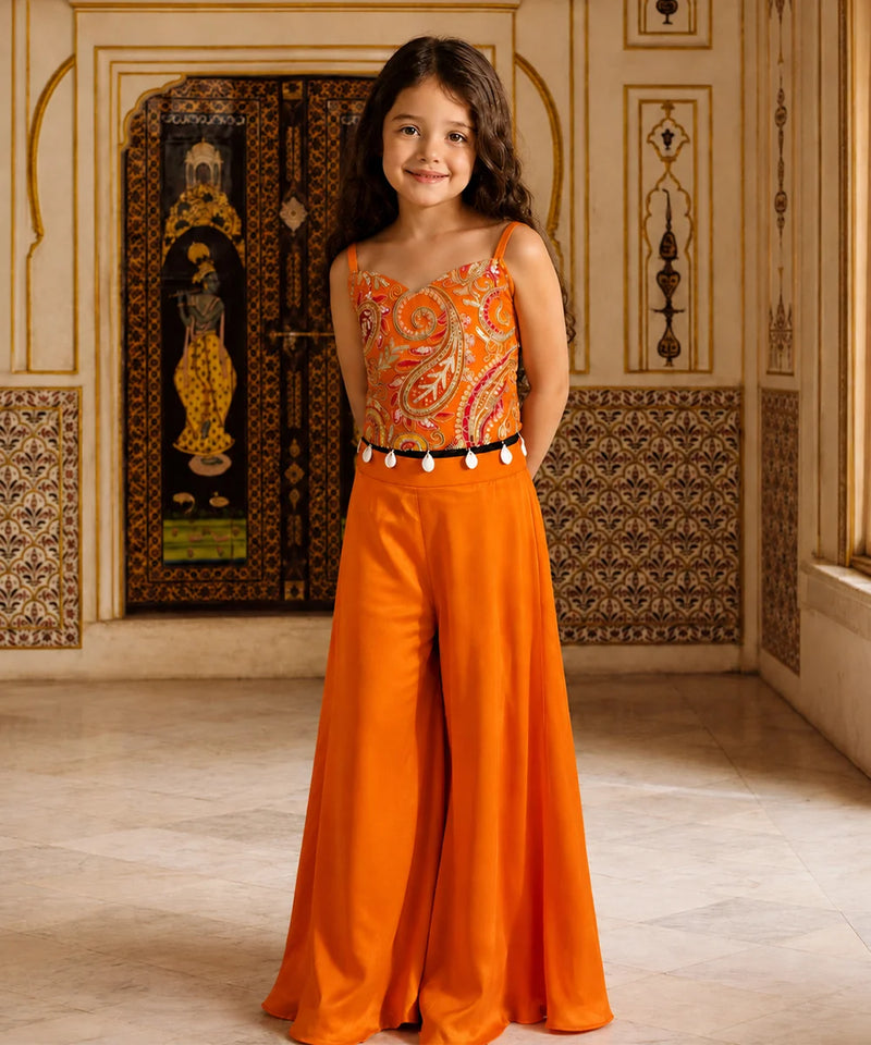 A young girl stands in an ornate indoor setting wearing a bright orange two-piece outfit with a paisley-patterned bodysuit top and matching wide-leg pants, accented by a black belt with white tassels.