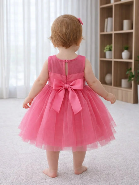 Child wearing a pink dress with a bow in a room with a bookshelf and plants.