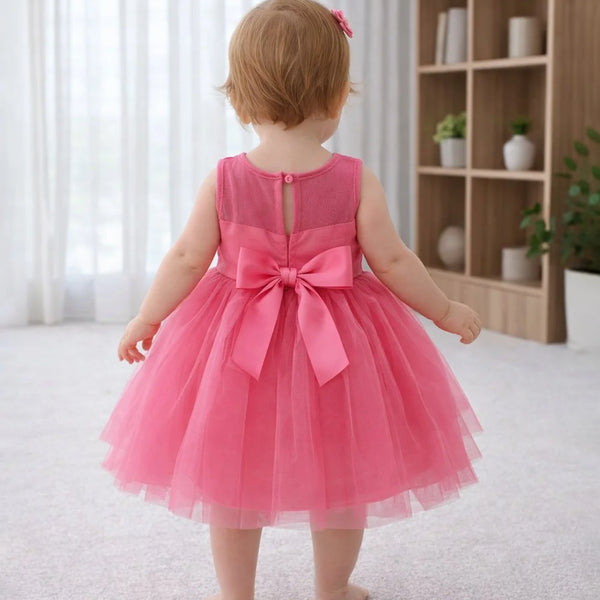Child wearing a pink dress with a bow in a room with a bookshelf and plants.