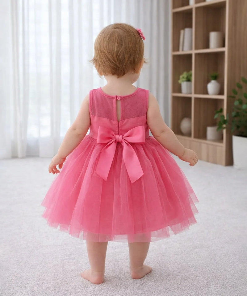 Child wearing a pink dress with a bow in a room with a bookshelf and plants.