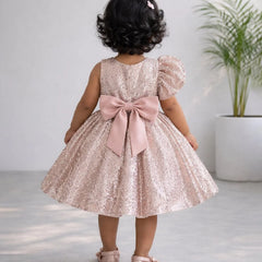 Child wearing a pink sequin dress with a large bow, standing indoors.