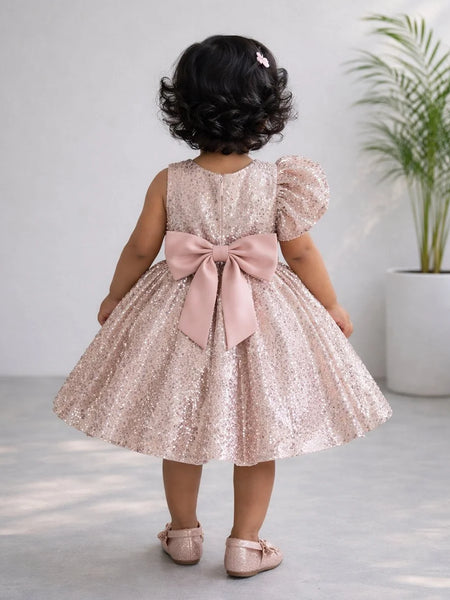 Child wearing a pink sequin dress with a large bow, standing indoors.