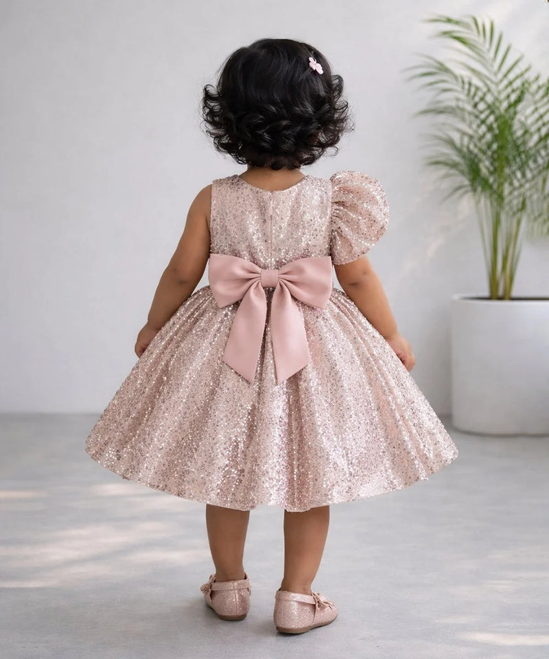 Child wearing a pink sequin dress with a large bow, standing indoors.