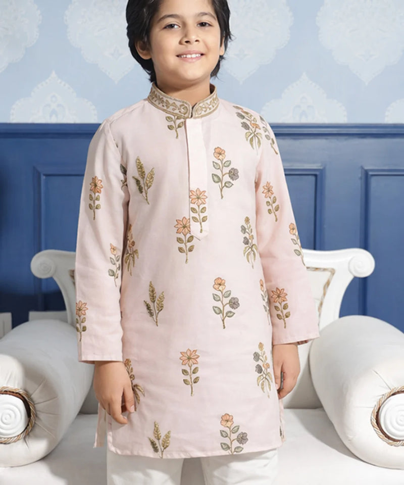 Child wearing a light pink embroidered kurta with white pants against a blue wall.