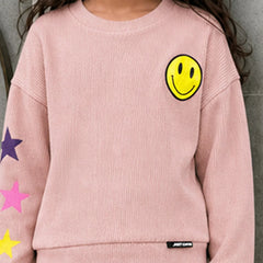 A child wearing a light pink ribbed knit sweatshirt featuring a yellow smiley face on the left chest, colorful star graphics on the sleeve, and a small black brand tag near the hem.