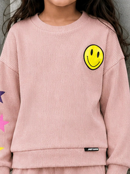 A child wearing a light pink ribbed knit sweatshirt featuring a yellow smiley face on the left chest, colorful star graphics on the sleeve, and a small black brand tag near the hem.