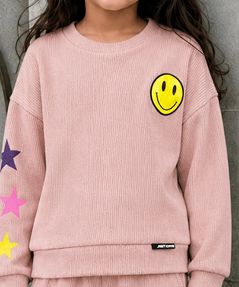 A child wearing a light pink ribbed knit sweatshirt featuring a yellow smiley face on the left chest, colorful star graphics on the sleeve, and a small black brand tag near the hem.
