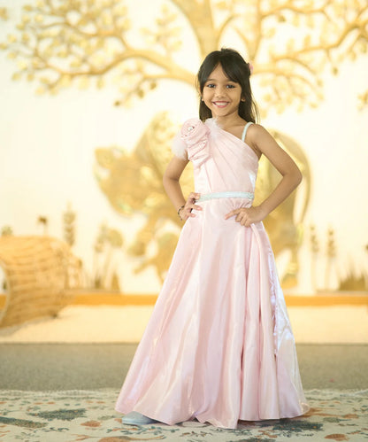 A young girl standing in a beautiful baby pink colored gown with a floral brooch on the shoulder and a belt detail on the waist, posing against a backdrop with a tree and decorative wall.