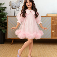 A young girl with long wavy brown hair is smiling and posing in a light pink tulle dress with a ruffled tuxedo-style collar and matching pink shoes, standing in a decorated room with a wooden cabinet and a horse statue in the background.