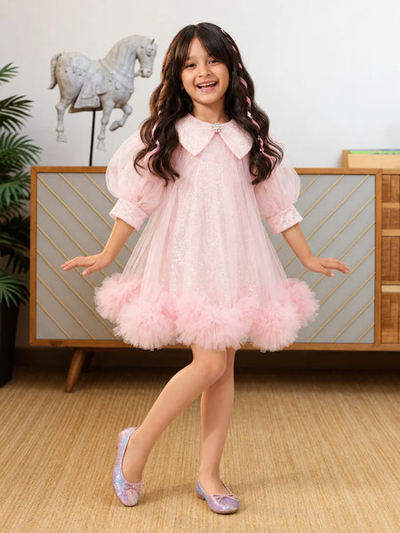 A young girl with long wavy brown hair is smiling and posing in a light pink tulle dress with a ruffled tuxedo-style collar and matching pink shoes, standing in a decorated room with a wooden cabinet and a horse statue in the background.