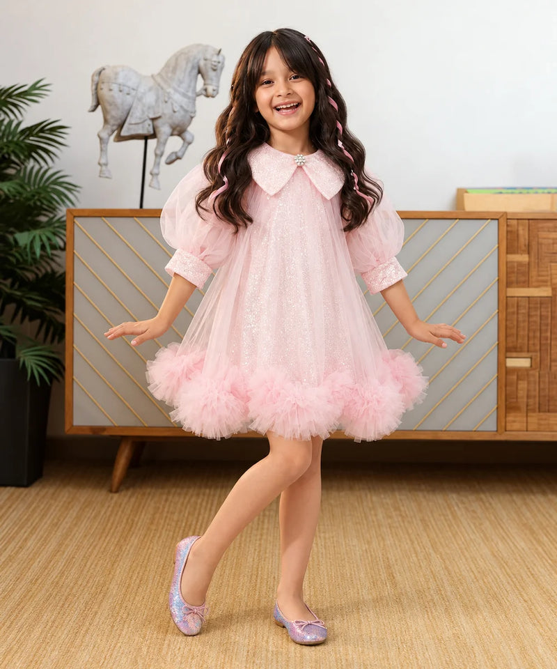 A young girl with long wavy brown hair is smiling and posing in a light pink tulle dress with a ruffled tuxedo-style collar and matching pink shoes, standing in a decorated room with a wooden cabinet and a horse statue in the background.